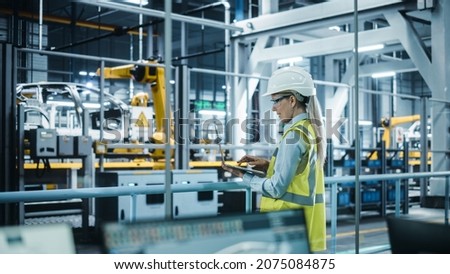 Car Factory: Female Automotive Engineer Wearing Hard Hat, Standing, Using Laptop. Monitoring, Control, Equipment Production. Automated Robot Arm Assembly Line Manufacturing Electric Vehicles. Royalty-Free Stock Photo #2075084875