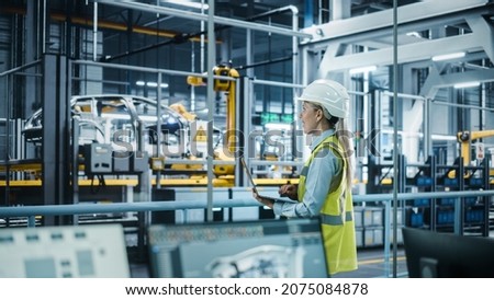 Car Factory: Female Automotive Engineer Wearing Hard Hat, Standing, Using Laptop. Monitoring, Control, Equipment Production. Automated Robot Arm Assembly Line Manufacturing Electric Vehicles. Royalty-Free Stock Photo #2075084878