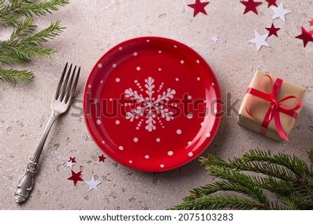 Red plate with white polka dots and snowflake with gift in craft paper tied with red ribbon decorated with Christmas spruce branches and confetti stars on alight beige background. Christmas background