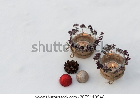 Against the background of snow, there are two candlesticks with burning candles. Glass candlesticks are decorated with oregano sprigs. The Christmas composition is complemented by cones and balls.