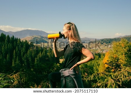 A profile picture of a young beautiful woman with long straight blonde hair, wearing black t-shirt, drinking from yellow thermo bottle, gorgeous landscape of mountains covered with coniferous forest