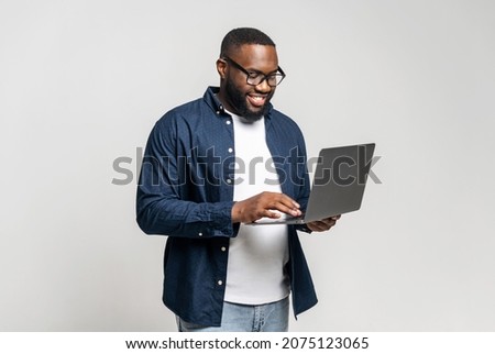 Portrait of young African American guy standing isolated on grey wall holding laptop, typing messages, chatting with friend on social media online, watching video, enjoying new application, websurfing Royalty-Free Stock Photo #2075123065