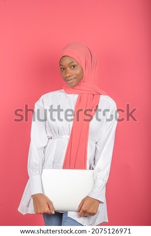 Image of an african american woman wearing pink scarf and holding a laptop isolated over pink background