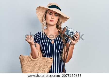 Beautiful young blonde woman holding vintage camera relaxed with serious expression on face. simple and natural looking at the camera. 