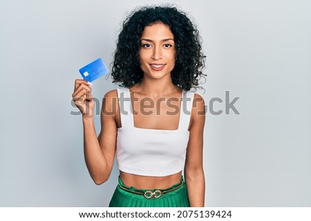 Young latin girl holding credit card looking positive and happy standing and smiling with a confident smile showing teeth 