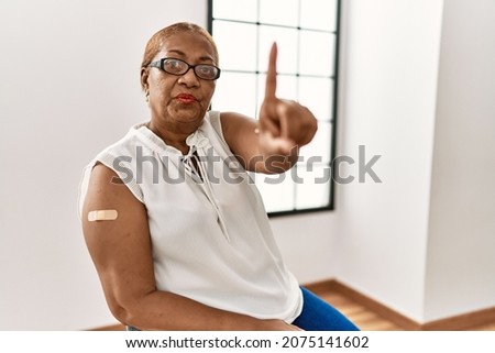 Mature hispanic woman getting vaccine showing arm with band aid pointing with finger up and angry expression, showing no gesture 
