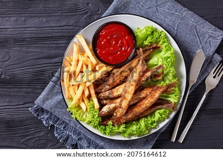 crispy fried capelin fish with fresh lettuce, french fries and tomato sauce on a plate on a black textured wooden table, flat lay, free space