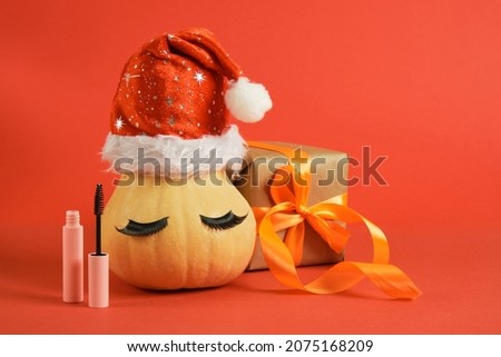 mascara in pink mock-up packaging, wrapped christmas gifts and pumpkin in santa hat and false eyelashes on a red background