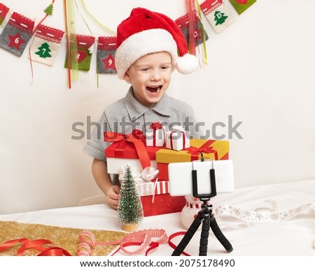 Happy little boy in red Santa Claus hat with Christmas gift boxes greets his friends and relatives by video call using smartphone. Christmas online greetings.