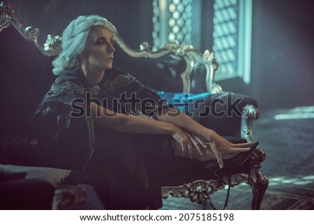 A beautiful old woman, a noble widow with gray hair and a rich black dress, sits on a vintage sofa in her old castle. Black widow. Fantasy world. Halloween.