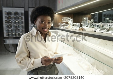 Young successful shop assistant holding tablet and looking at you while standing by display with jewelry Royalty-Free Stock Photo #2075205160