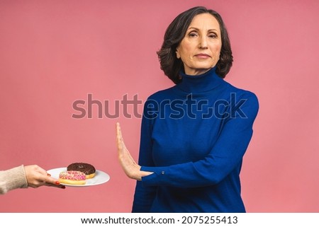 Stop sign. Charming happy aged mature senior woman refused to eat glazed donut dressed in casual isolated over pink background. Old age and diet concept.