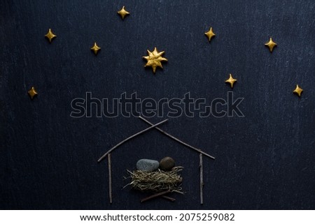 a figurine of a baby folded of stones, symbolizing Jesus Christ, lying in a manger in a small house made of tree branches, under a starry sky with a Christmas star on a dark stone surface