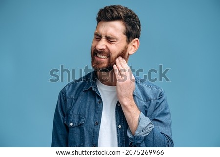 Dental problems. Portrait of unhealthy man pressing sore cheek, suffering acute toothache, periodontal disease, cavities or jaw pain. Indoor studio shot isolated on blue background Royalty-Free Stock Photo #2075269966