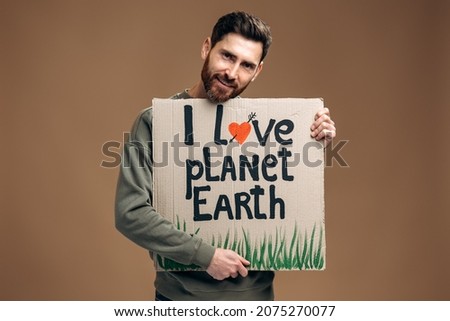 Caucasian brunette bearded man holding carton placard with I love planet earth and embracing it with pleasure smile. Concept of eco activism