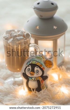 Cozy winter holiday morning breakfast with cute penguin gingerbread cookie and coffee or cocoa with marshmallow. Christmas inspiration. Cup of hot chocolate with candle in lantern and garland lights