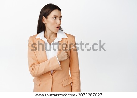 Surprised corporate woman pointing and looking right at logo, showing advertisement aside, standing in business suit over white background