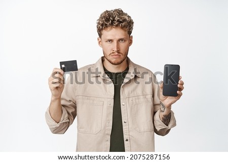 Angry young man shows credit card and mobile phone screen, not enough fonds on bank account, showing mobile banking app without money, white background