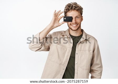 Happy young man smiles, shows his credit card near face, concept of money and shopping, stands against white background