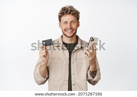 Handsome smiling blond male model, showing credit card and holding smartphone, paying online, shopping in internet store, white background