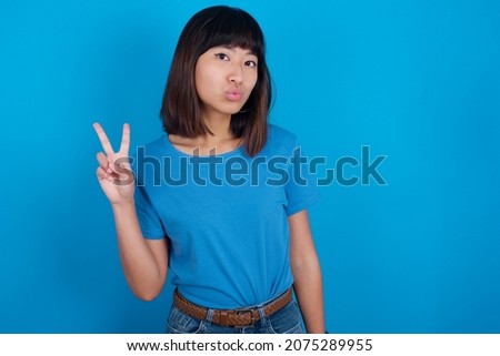 young asian woman wearing blue t-shirt against blue background makes peace gesture keeps lips folded shows v sign. Body language concept
