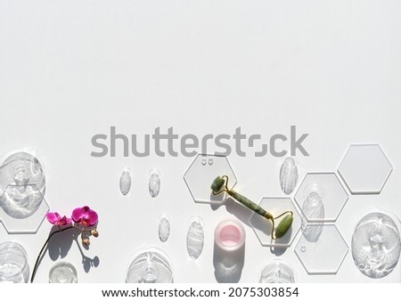 Jade stone face roller with fuchsia orchid flowers and glass baubles. Pink moisturizer cream in jar. Sunshine, long shadows. Flat lay on off white background with copy-space. Face lymph drainage.