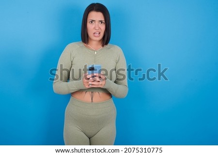 Photo of astonished crazy fitness caucasian sporty girl standing against blue background hold smartphone dislike feedback concept