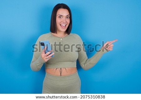 Astonished fitness caucasian sporty girl standing against blue background holding her telephone and pointing with finger aside at empty copy space