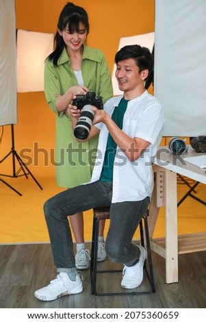 Happy professional male photographer holding DSLR camera smiling while showing pictures on camera screen to pretty young model who feels surprised with shy in what she sees.