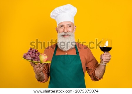 Portrait of attractive bearded cheerful grey-haired man holding in hand wine snack treat isolated over bright yellow color background