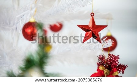 Close up shot of glossy shiny red Xmas tree shape decorating object hanged on white leaves decoration Christmas tree branch with other star ball and gold ribbon present gift box in blurred background.