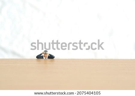 Businessman using binoculars hiding on wooden desk. Miniature tiny people toys photography. isolated on white background.