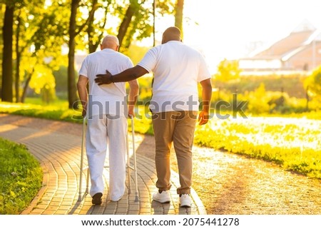 Caregiver is teaching old man to walk with walker. Professional nurse and patient walking outdoor at sunset. Assistance, rehabilitation and health care. Royalty-Free Stock Photo #2075441278