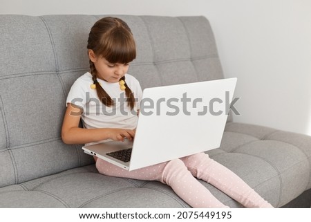 Dark haired charming adorable little girl with pigtail sitting on sofa in light living room and holding notebook, writing on keyboard or watching cartoon or videos.