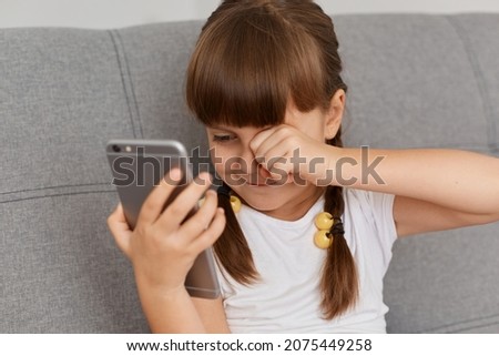 Indoor shot of tired cute female kid wearing casual style clothing posing in light room, using smart phone long hours and suffering eyes hurt, rubbing her eyes, looks sleepy.