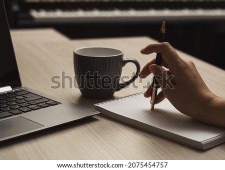 Close-up woman's hand holding a pencil and writing data in a notebook next to a laptop and cup of coffee on a beige wood table against the background of a piano keyboard. Horizontal view.