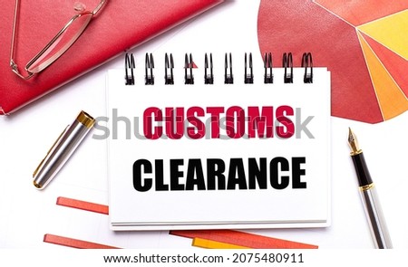 On the desktop is a white notebook with the text CUSTOMS CLEARANCE, a pen, burgundy and red tables, and gold-framed glasses. Business concept.