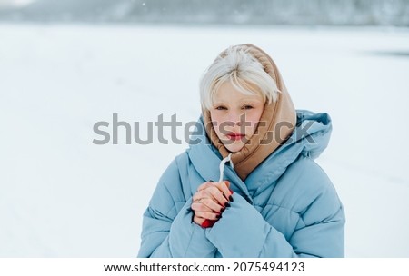 Frozen blonde girl stands on a winter street and looks at the camera with a serious face on a snowy background in the snowfall.
