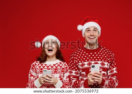 Young fun couple friends two man woman in sweater hat hold in hand use mobile cell phone look overhead isolated on plain red background. Happy New Year 2022 celebration merry ho x-mas holiday concept