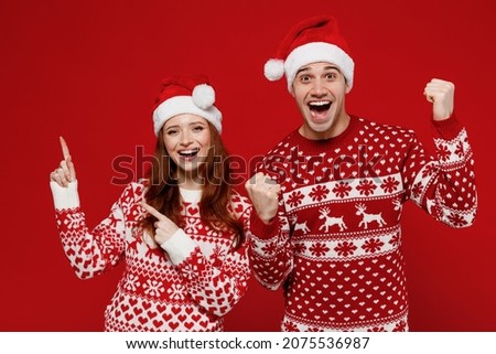 Young couple friends two man woman 20s in sweater hat point index finger aside on copy space workspace area do winner gesture isolated on plain red background. Happy New Year 2022 celebration concept