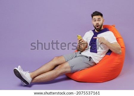 Full size body surprised fun young brunet man 20s wear white t-shirt purple shirt sit in bag chair hold in hand use point on mobile cell phone show isolated on pastel violet background studio portrait