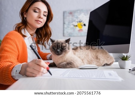 young woman signing blurred contract near monitor with blank screen and cat on desk