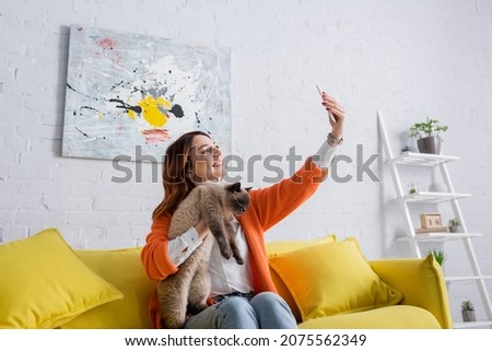 excited woman taking selfie with cat while sitting on couch near picture on white wall