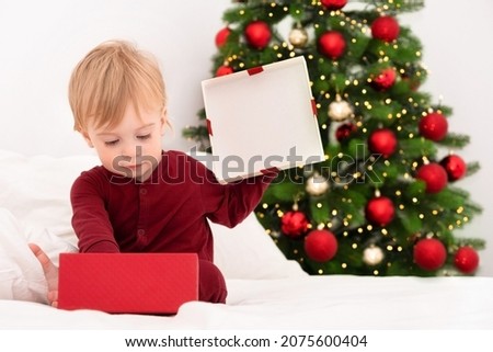Cute little blonde caucasian baby get red gift box by Christmas tree in bedroom.Spend winter holiday.Giving New Year present to infant,kid,toddler.Greeting card,mock-up,copy space.Selective focus