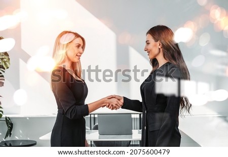 Businesswomen in formal wear shake hands in light office room with desk and laptop, bokeh lights. Happy and smiling business partners working together. Concept of financial deal