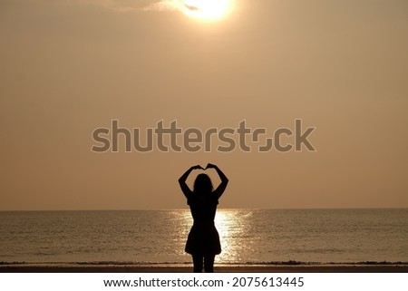 The girl's back shadow made a heart shape with her hands on her head. In the background of the sunset, orange light reflects against the evening sea.