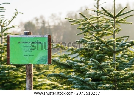 Sign with German text "Christmas trees Saturday, 19.12.2020, 10:00-12:30, Monday, 21.12.2020, 13:00-16:00 or by appointment" in front of frost covered evergreen fir trees on a Christmas tree farm.