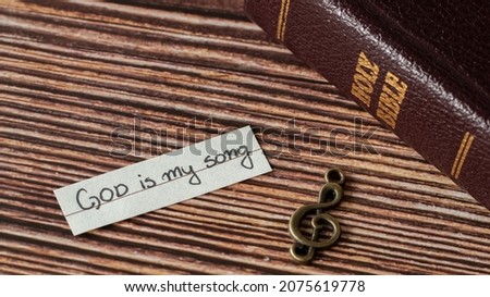 A rustic old treble clef note, handwritten quote: God is my song, and closed Holy Bible on wooden table. Christian biblical concept of music, praise, worship, and joy in the LORD. A closeup. Royalty-Free Stock Photo #2075619778