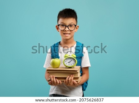 Time To Study Concept. Little Asian Schoolboy Holding Stack Of Books, Green Apple And Clock While Posing Over Blue Studio Background, Nerdy Korean Kid With Backpack Enjoying Learning, Copy Space