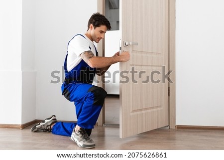 Installation of a lock on the front wooden entrance door. Portrait of young locksmith workman in blue uniform installing door knob. Professional repair service. Maintenance Concept Royalty-Free Stock Photo #2075626861